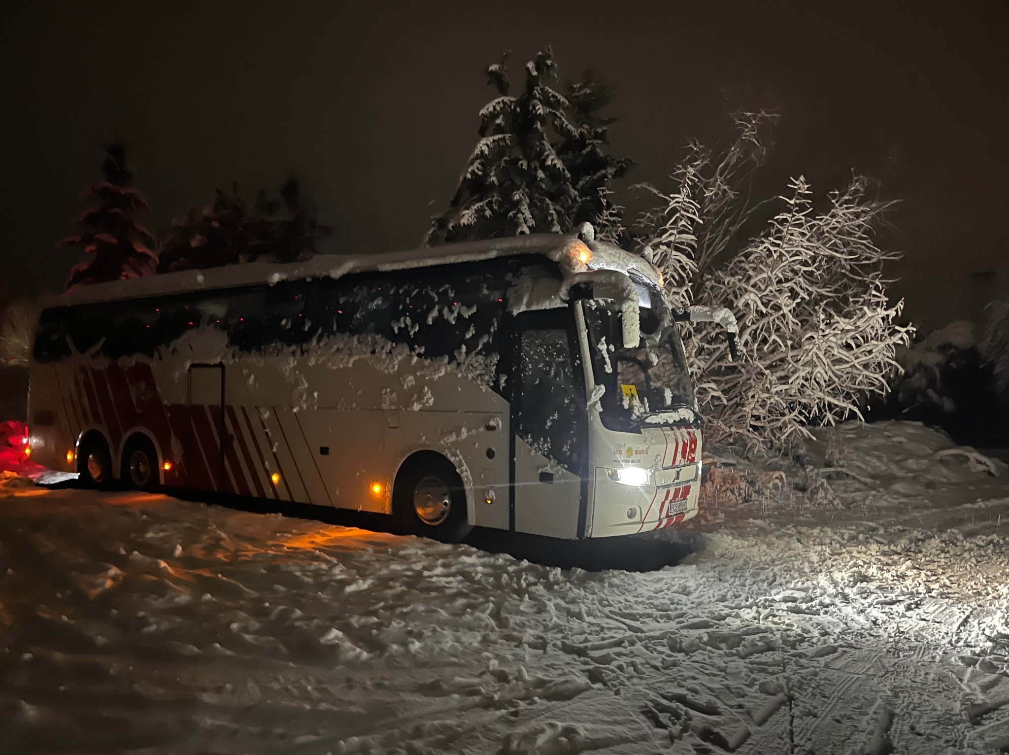 Autokar w zimowej scenerii nocą, ośnieżony dach i drzewa. Widoczne światła pojazdu oświetlają śnieg. Czerwone pasy na karoserii.