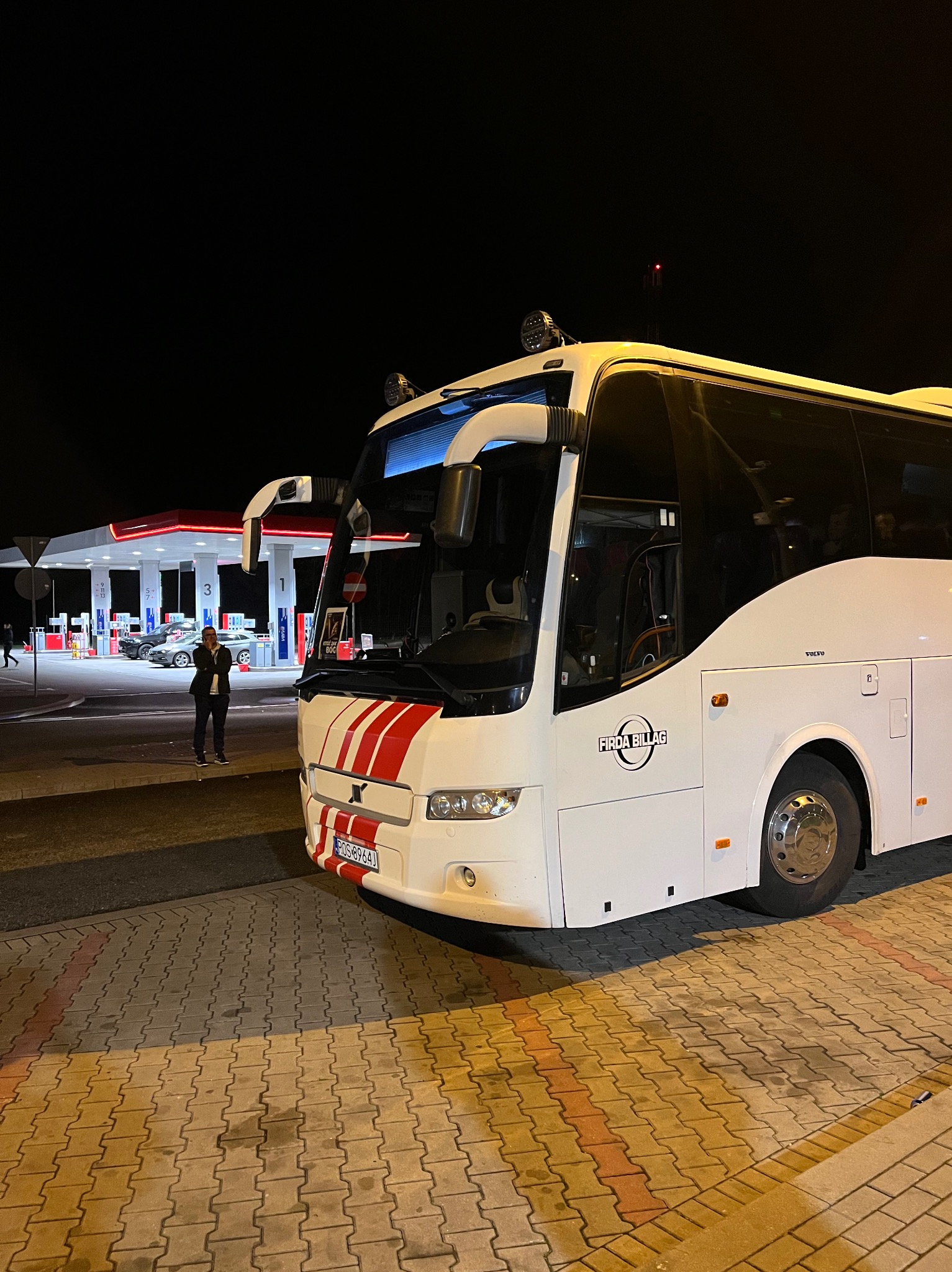 Biały autokar Volvo z czerwonymi pasami na tle stacji benzynowej nocą. Widoczny mężczyzna rozmawiający przez telefon i logo firmy 'Firda Billag'.