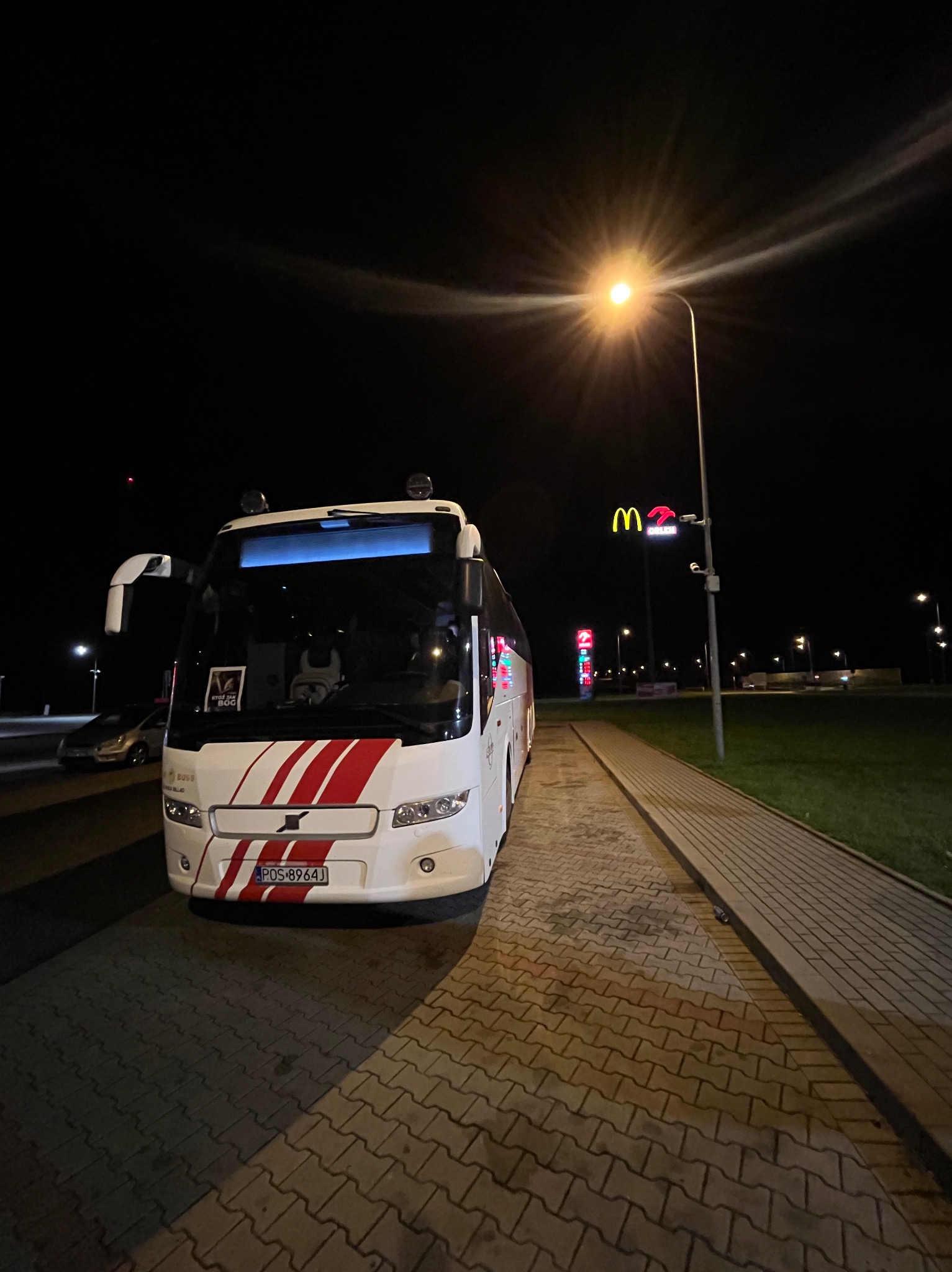 Biały autokar z czerwonymi pasami parkuje nocą przy oświetlonej ulicy, w tle widoczny neon McDonald's. Tablica rejestracyjna widoczna.