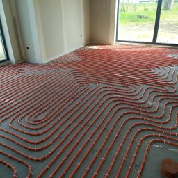 Filar DOM sp. z o.o. sp.k. - Pomarańczowe rury ogrzewania podłogowego, równolegle ułożone na szarej folii w jasnym pomieszczeniu z dużymi oknami, widok z góry, etap instalacji.