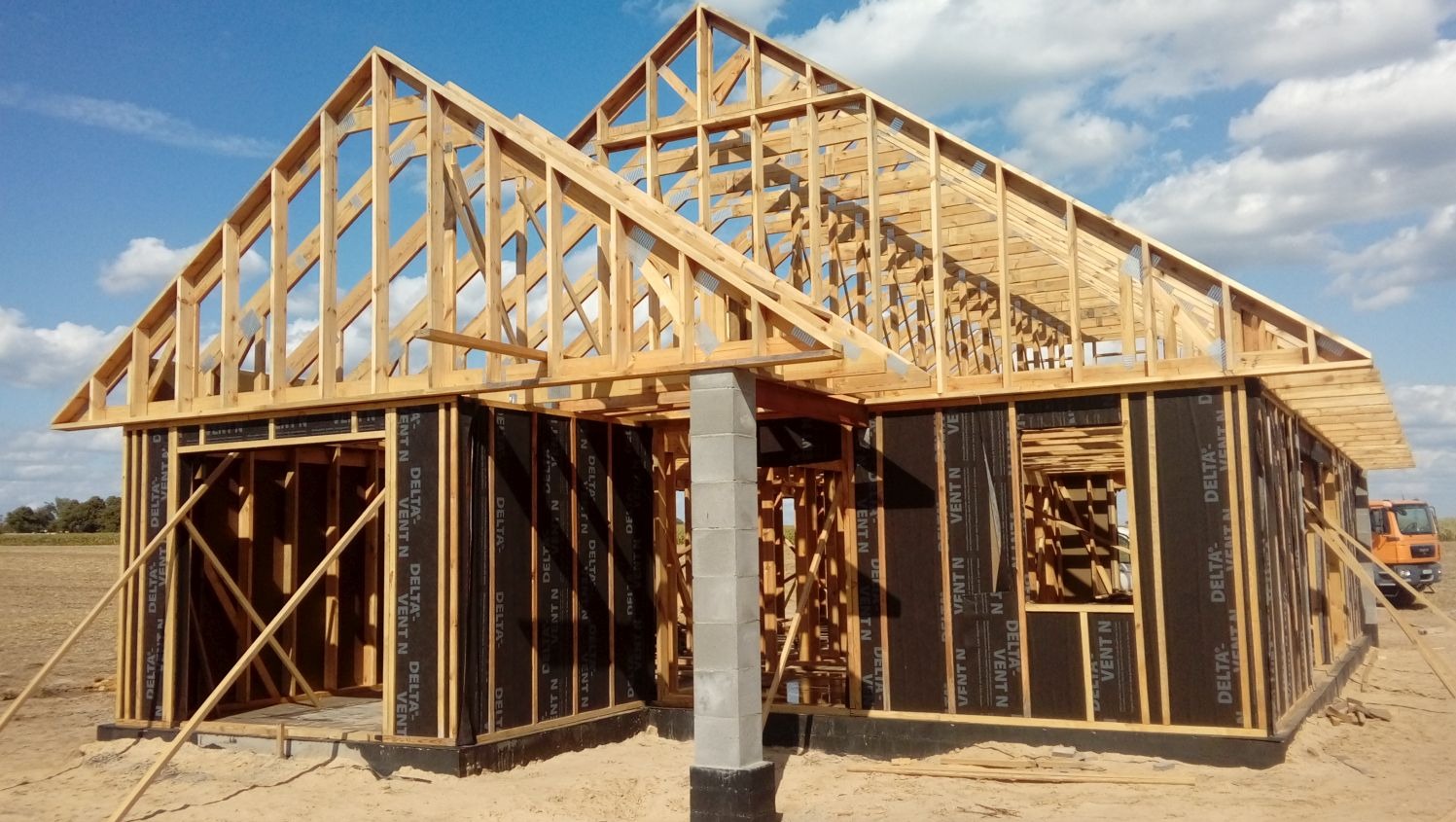 Konstrukcja domu w trakcie budowy: drewniany szkielet z czarną membraną, betonowy filar, błękitne niebo. Widoczny fragment pomarańczowej ciężarówki w tle.