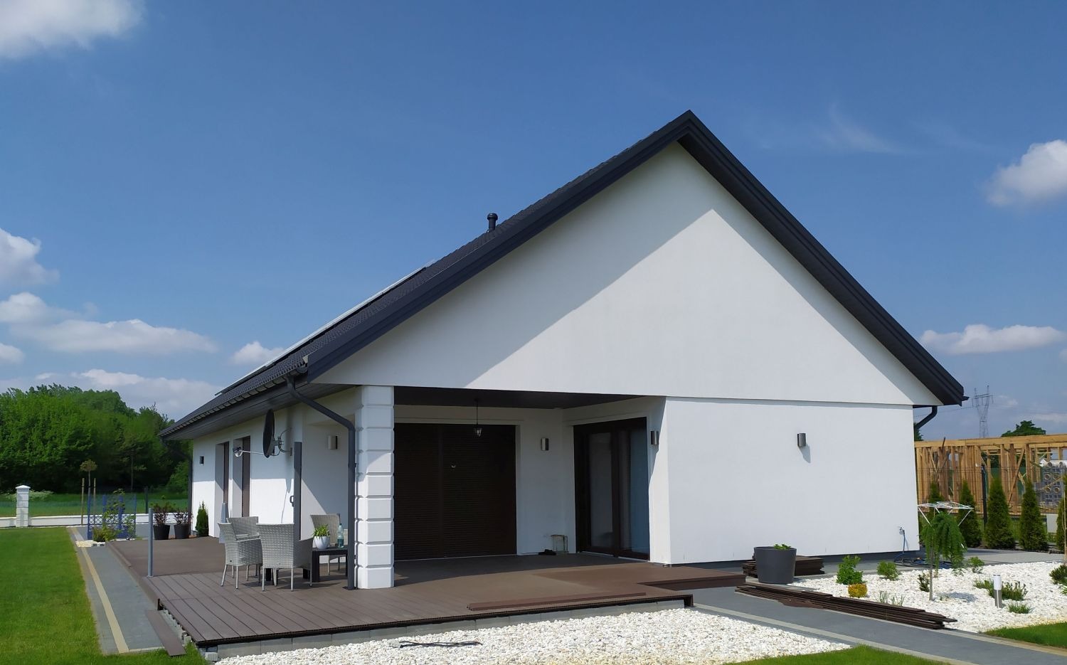 Nowoczesny, parterowy dom z białymi ścianami i ciemnym dachem, otoczony zielonym trawnikiem i dekoracyjnym żwirem. Widoczny taras z meblami ogrodowymi.