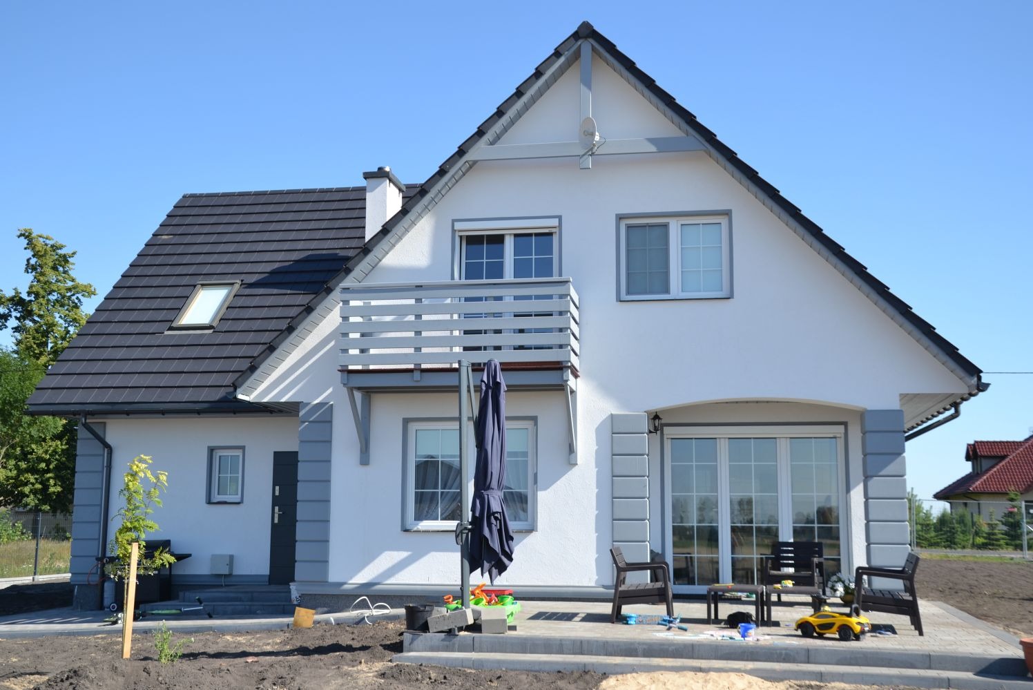 Nowoczesny dom z balkonem i tarasem, biała elewacja z szarymi akcentami, ciemna dachówka, zabawki na tarasie, świeżo zasadzona zieleń, błękitne niebo.