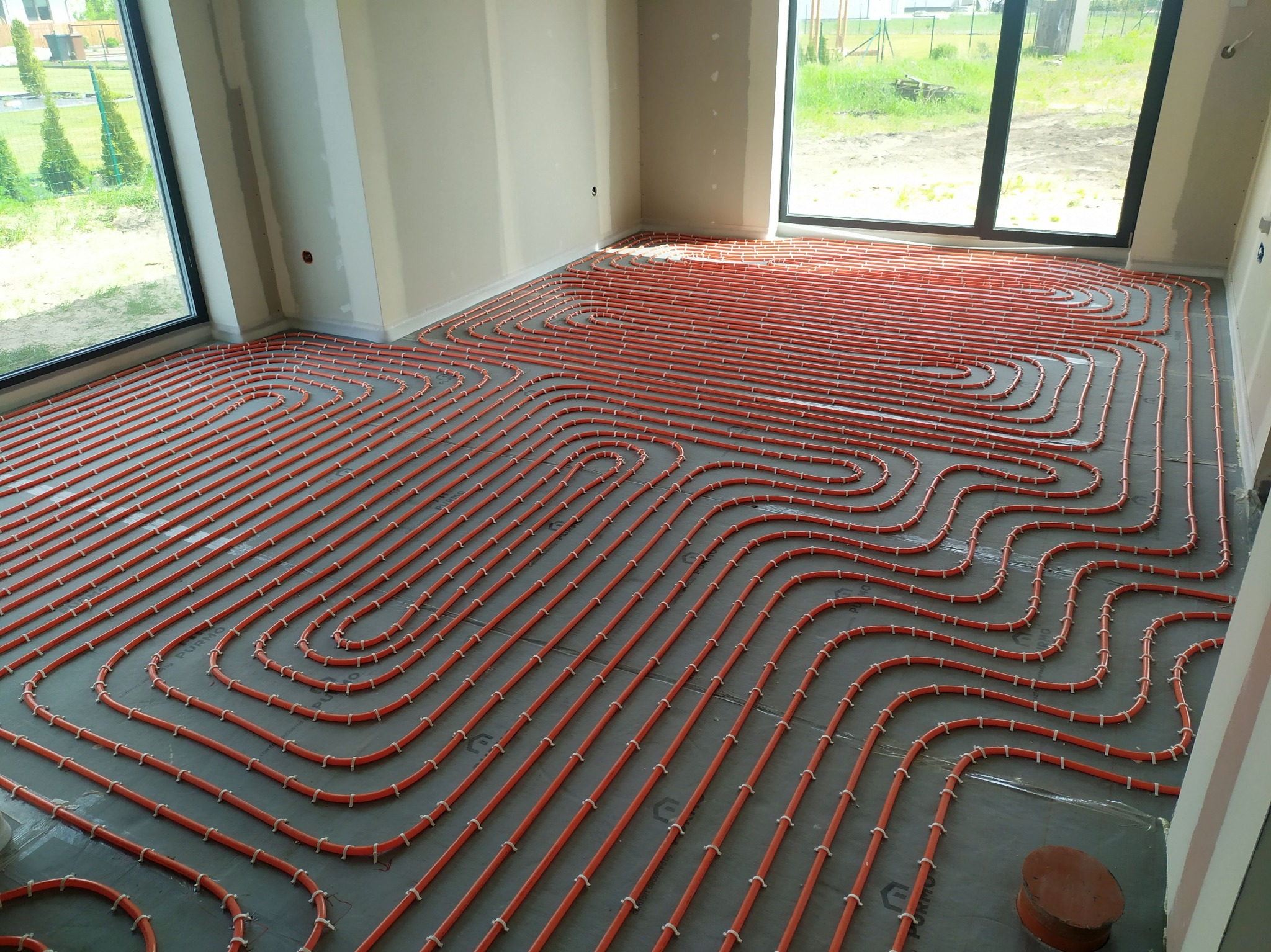 Pomarańczowe rury ogrzewania podłogowego, równolegle ułożone na szarej folii w jasnym pomieszczeniu z dużymi oknami, widok z góry, etap instalacji.