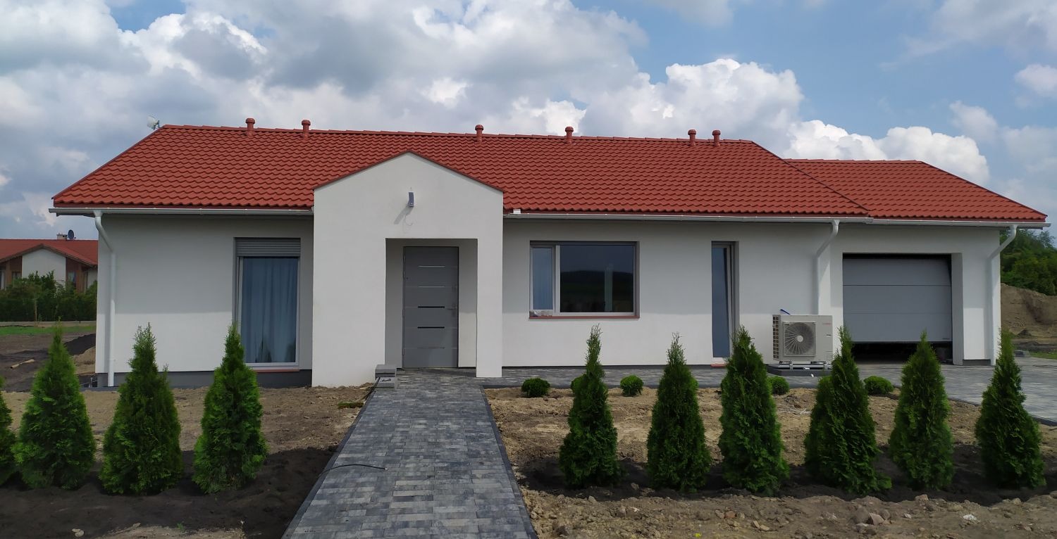 Parterowy dom z czerwoną dachówką, szarą stolarką i klimatyzatorem. Uwagę przykuwa ścieżka z kostki prowadząca do wejścia oraz rząd małych, zielonych drzewek.
