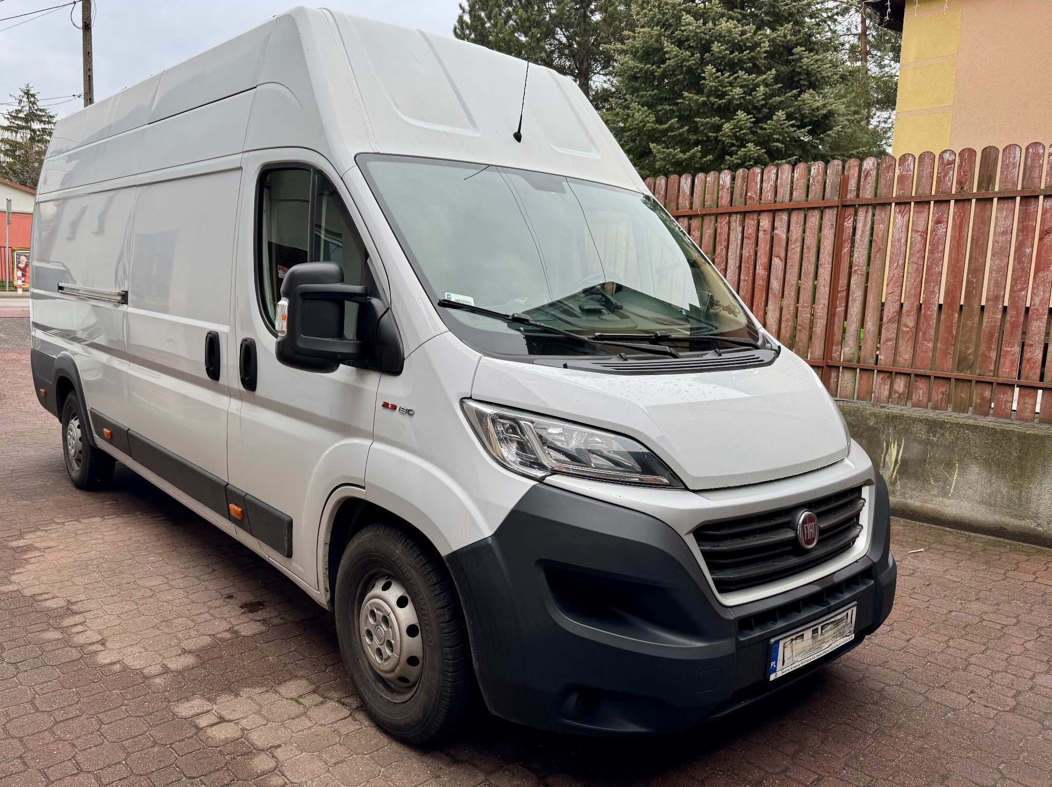 Biały bus Fiat Ducato z wysokim dachem, zaparkowany na brukowanej nawierzchni, z widocznym fragmentem drewnianego ogrodzenia w tle. Ujęcie z niskiej perspektywy.