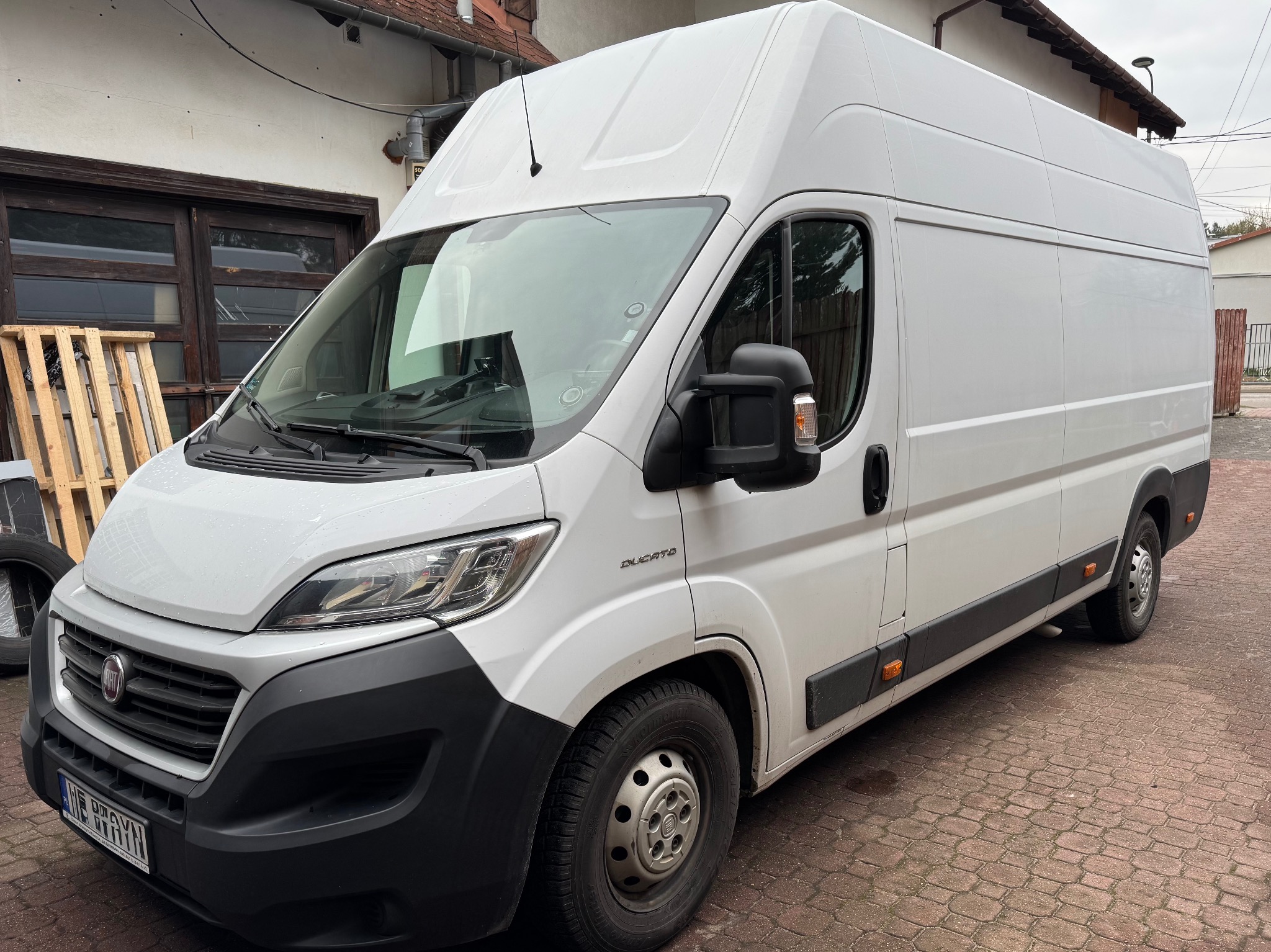 Biały furgon Ducato zaparkowany na brukowanej nawierzchni, widok z boku, z paletą w tle. Auto gotowe do transportu lub przeprowadzki.