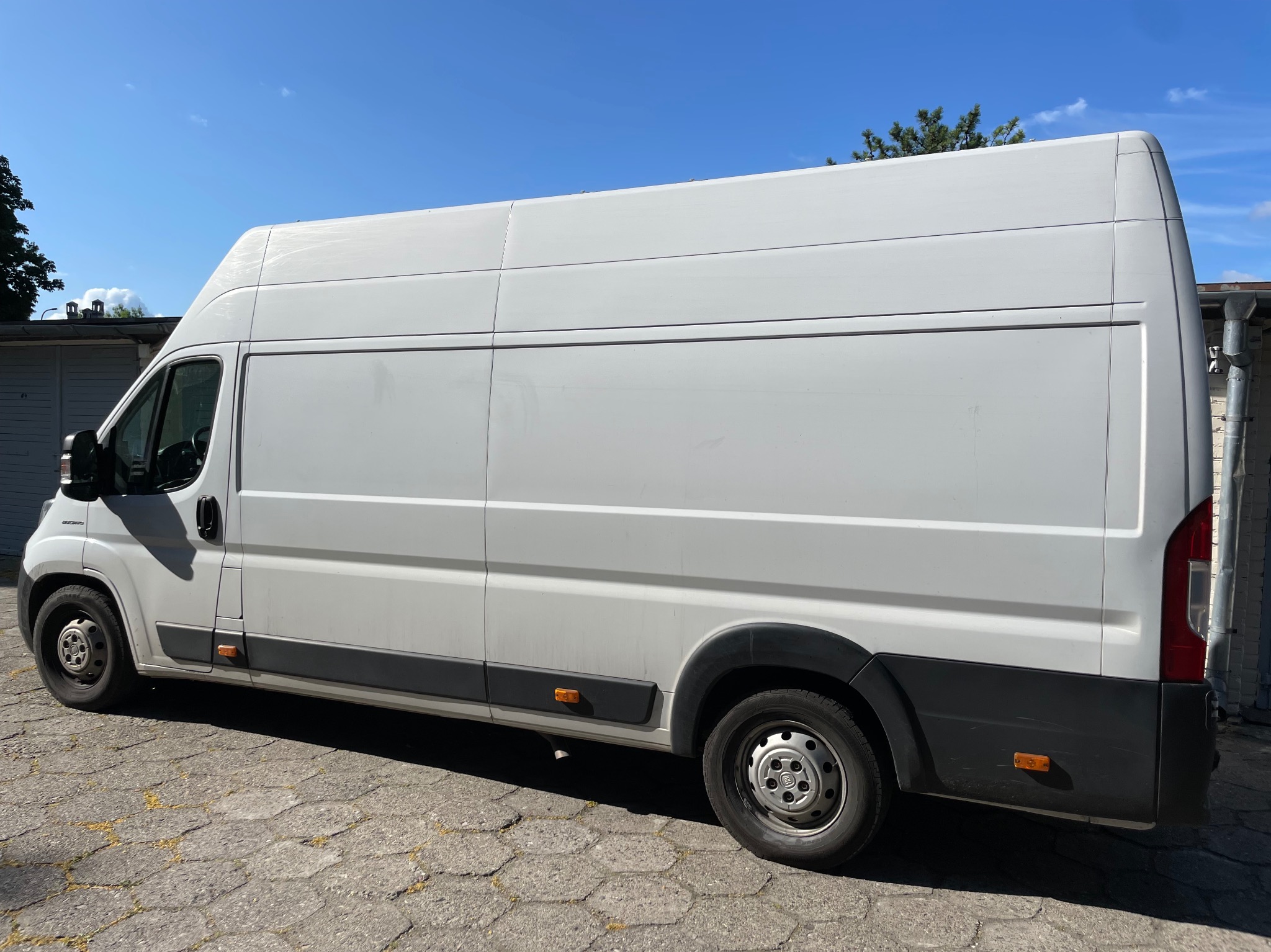 Biały, dostawczy bus marki Fiat Ducato, zaparkowany na brukowanej nawierzchni, z widocznymi detalami karoserii i opon. Błękitne niebo w tle.