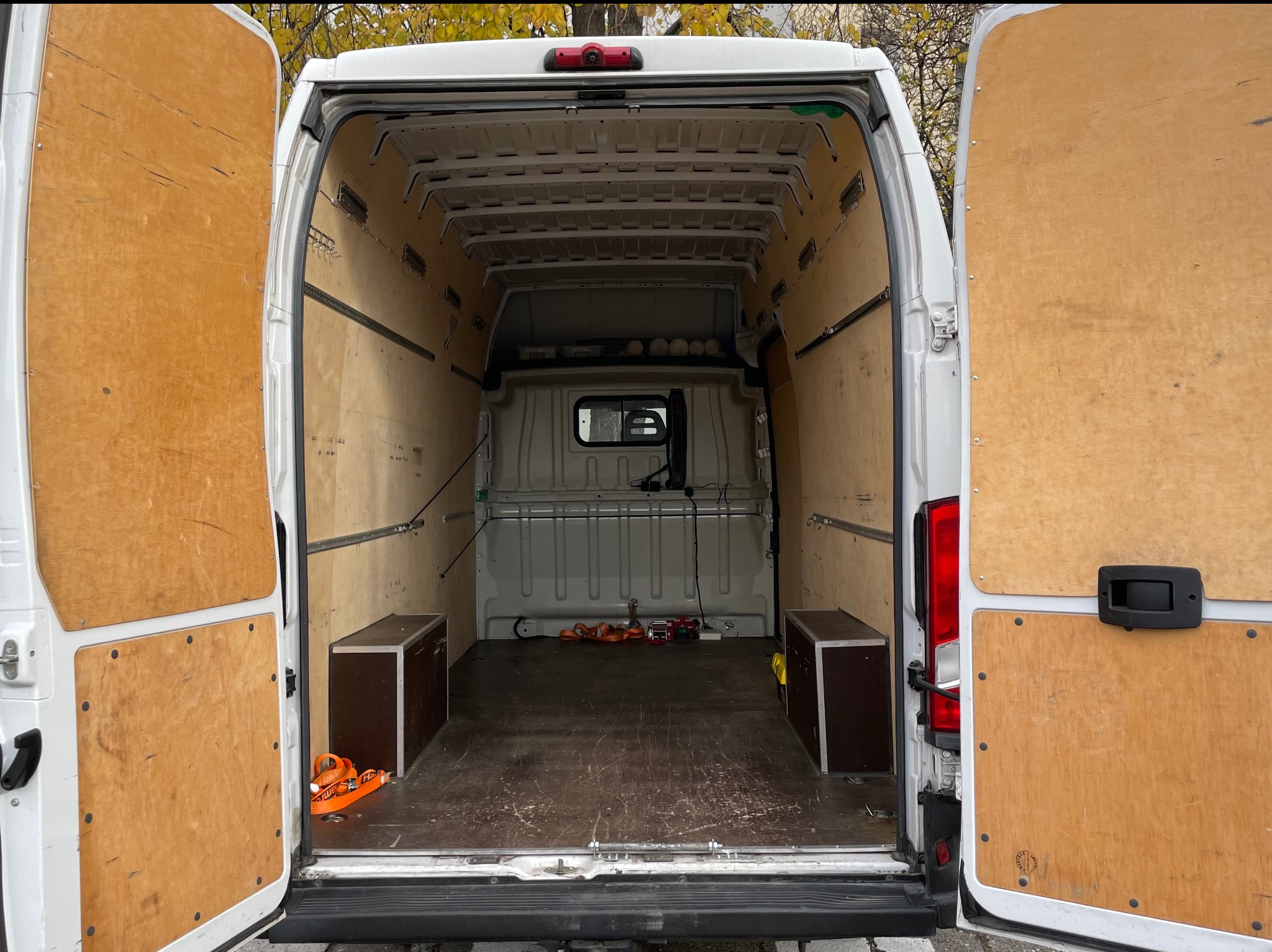 Wnętrze białego busa z otwartymi drzwiami, wyłożone sklejką, z dwoma skrzyniami i pasami transportowymi. Widok z tyłu pojazdu.