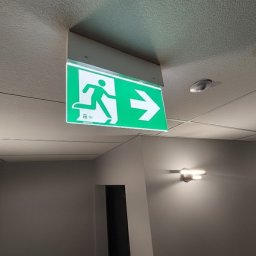 ZELITE Pomiary Projekty elektryczne - Zielony znak ewakuacyjny z piktogramem biegnącej osoby i strzałką w prawo, zamontowany na suficie w korytarzu z oświetleniem ściennym.