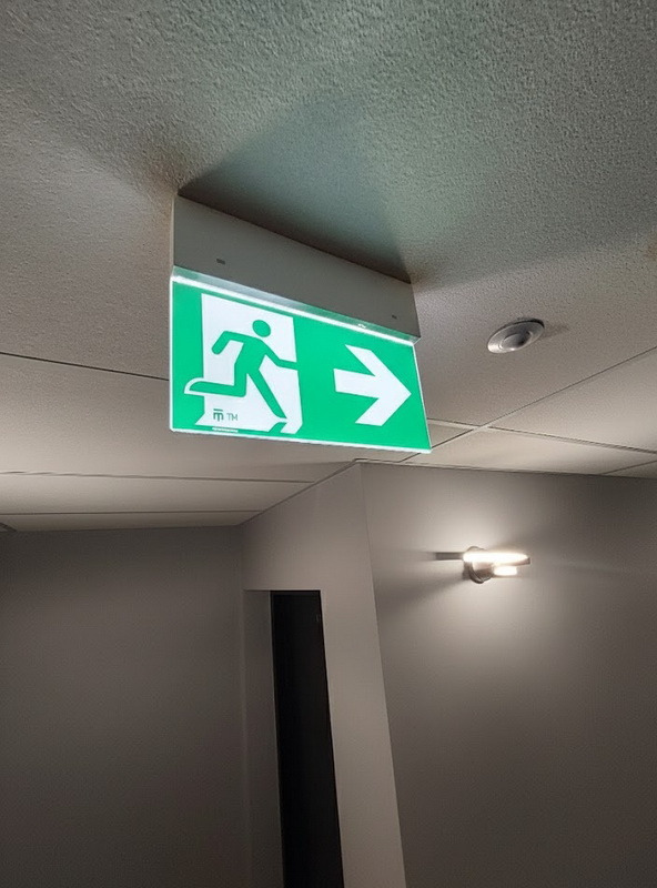 Zielony znak ewakuacyjny z piktogramem biegnącej osoby i strzałką w prawo, zamontowany na suficie w korytarzu z oświetleniem ściennym.