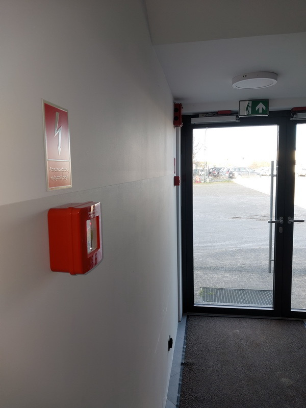 Korytarz z oznaczeniami bezpieczeństwa: znak ostrzegawczy wysokiego napięcia, czerwona skrzynka alarmowa, wyjście ewakuacyjne nad drzwiami. Widok na zewnątrz.