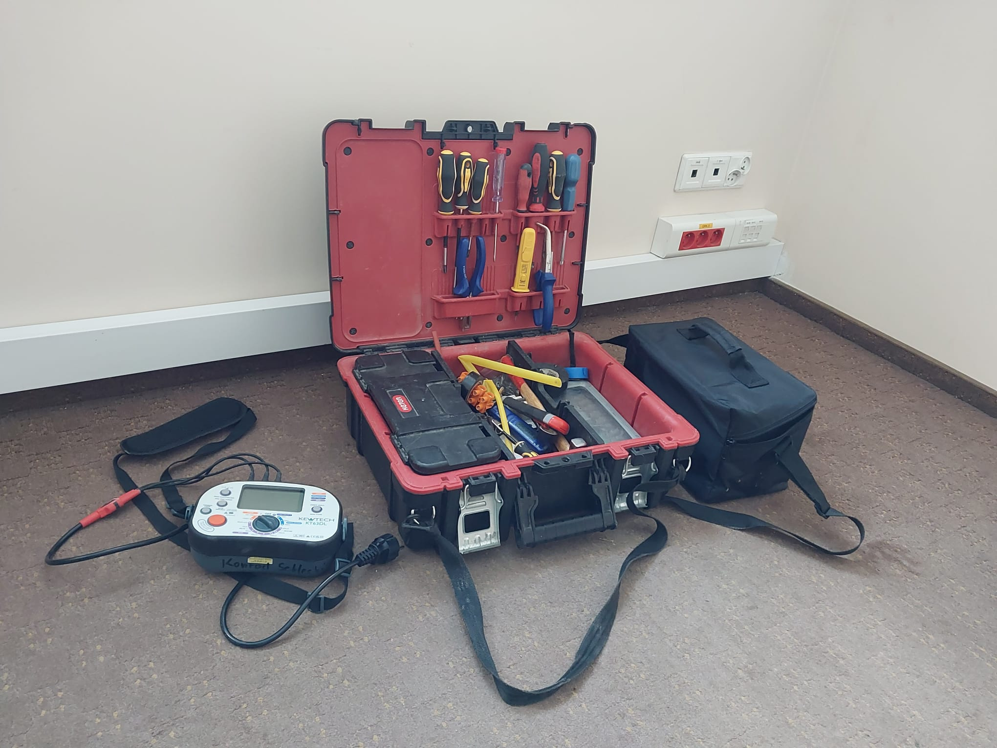 Otwarta skrzynka narzędziowa z miernikiem elektrycznym Kewtech KT63DL na podłodze, obok gniazd elektrycznych. Widoczne śrubokręty i inne narzędzia.