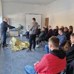 MULTI-SZKOL SPÓŁKA Z OGRANICZONĄ ODPOWIEDZIALNOŚCIĄ - Instruktor w czarnej bluzie demonstruje użycie koca termicznego na podłodze, podczas gdy grupa mężczyzn uważnie obserwuje w jasnej sali szkoleniowej.