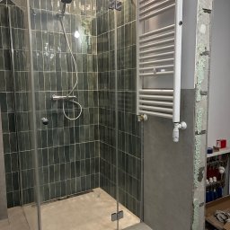 Ivan Vovk - Nowoczesna kabina prysznicowa z ciemnozielonymi, pionowo ułożonymi płytkami. Widoczny grzejnik drabinkowy i fragment ściany w trakcie prac wykończeniowych.