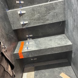 Ivan Vovk - Montaż grafitowych płytek na schodach z użyciem klipsów poziomujących. Widoczny fragment osoby w roboczej kurtce i fragment podłogi z płytką.