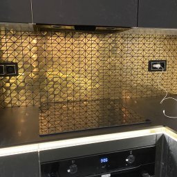 Ivan Vovk - Kuchnia z czarnymi szafkami i złotą mozaiką na ścianie nad blatem. Widoczne gniazdka elektryczne i płyta indukcyjna. Oświetlenie LED pod szafkami dodaje blasku.
