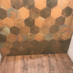 Ivan Vovk - Ściana w łazience wyłożona heksagonalnymi płytkami w odcieniach brązu, miedzi i turkusu, z drewnopodobną podłogą. Widoczny fragment półki i armatury.