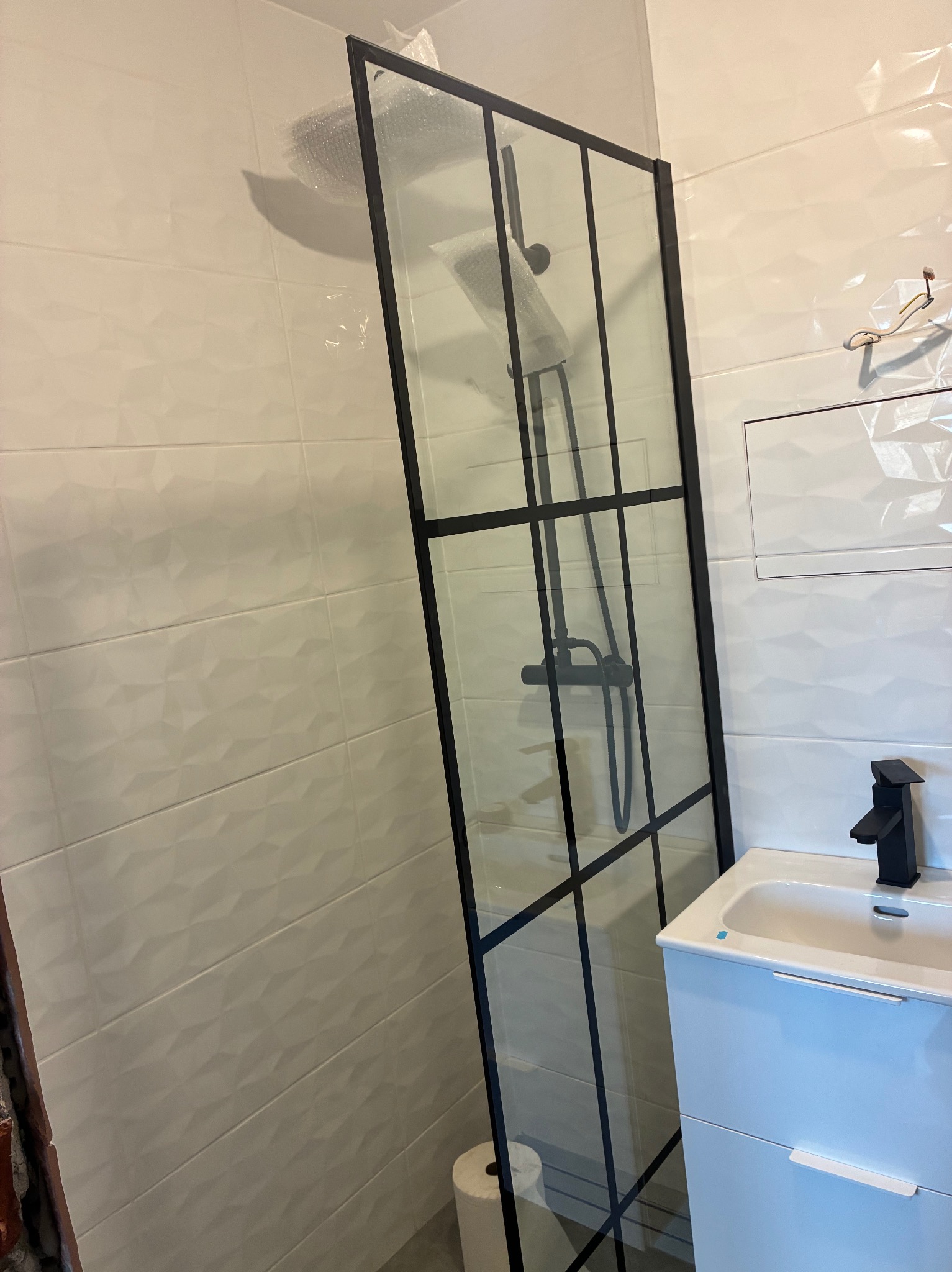 Nowoczesna kabina prysznicowa z czarnymi profilami na tle białych, trójwymiarowych płytek. Umywalka z czarną armaturą. Minimalistyczny design łazienki.