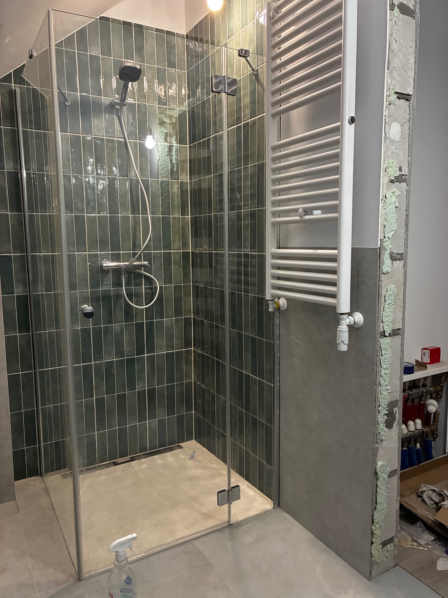 Nowoczesna kabina prysznicowa z ciemnozielonymi, pionowo ułożonymi płytkami. Widoczny grzejnik drabinkowy i fragment ściany w trakcie prac wykończeniowych.