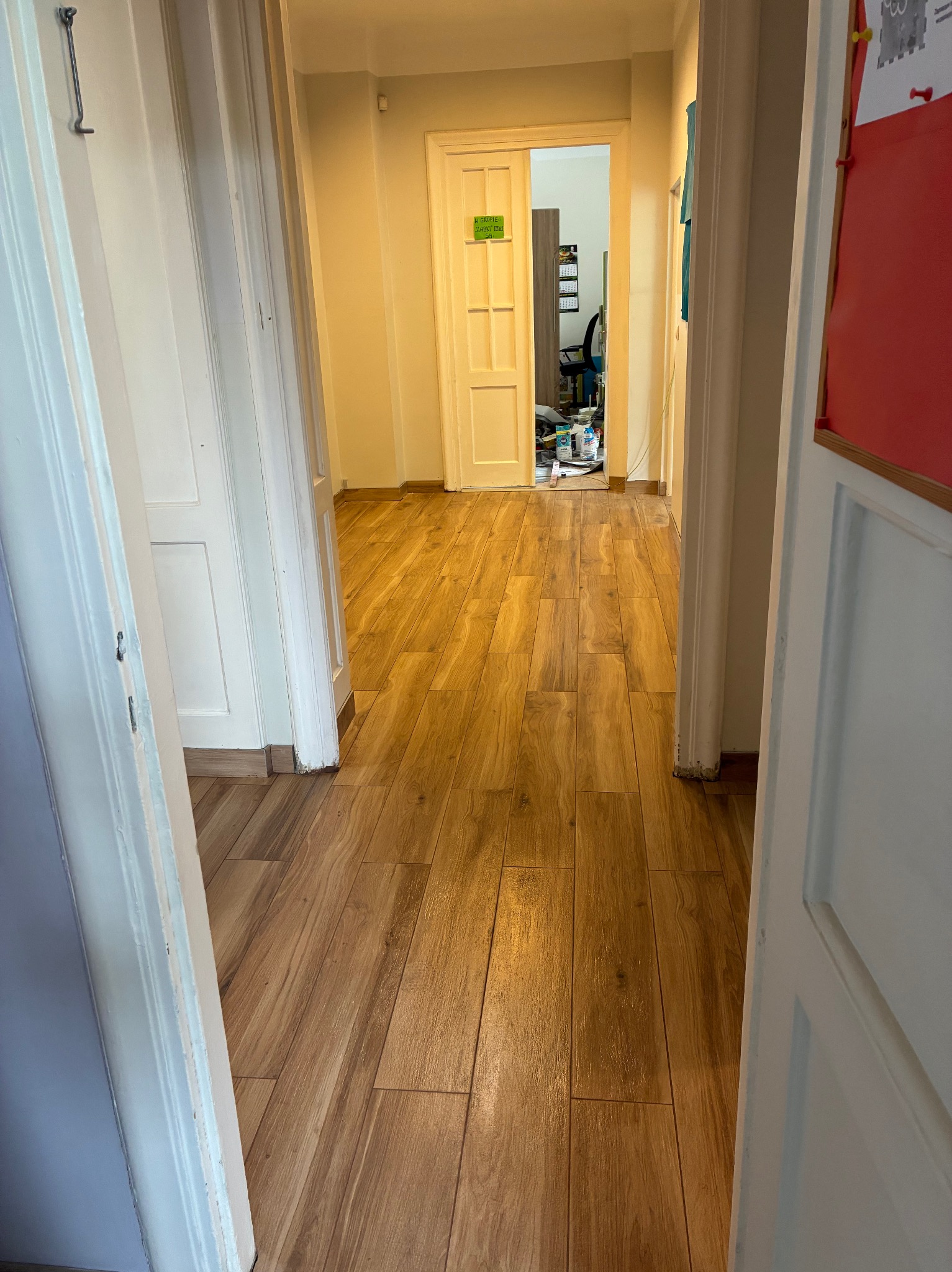 Ułożona podłoga z paneli imitujących drewno w korytarzu, widok przez otwarte drzwi. Widoczne białe drzwi i ściany w ciepłych odcieniach. W tle pomieszczenie z wyposażeniem.