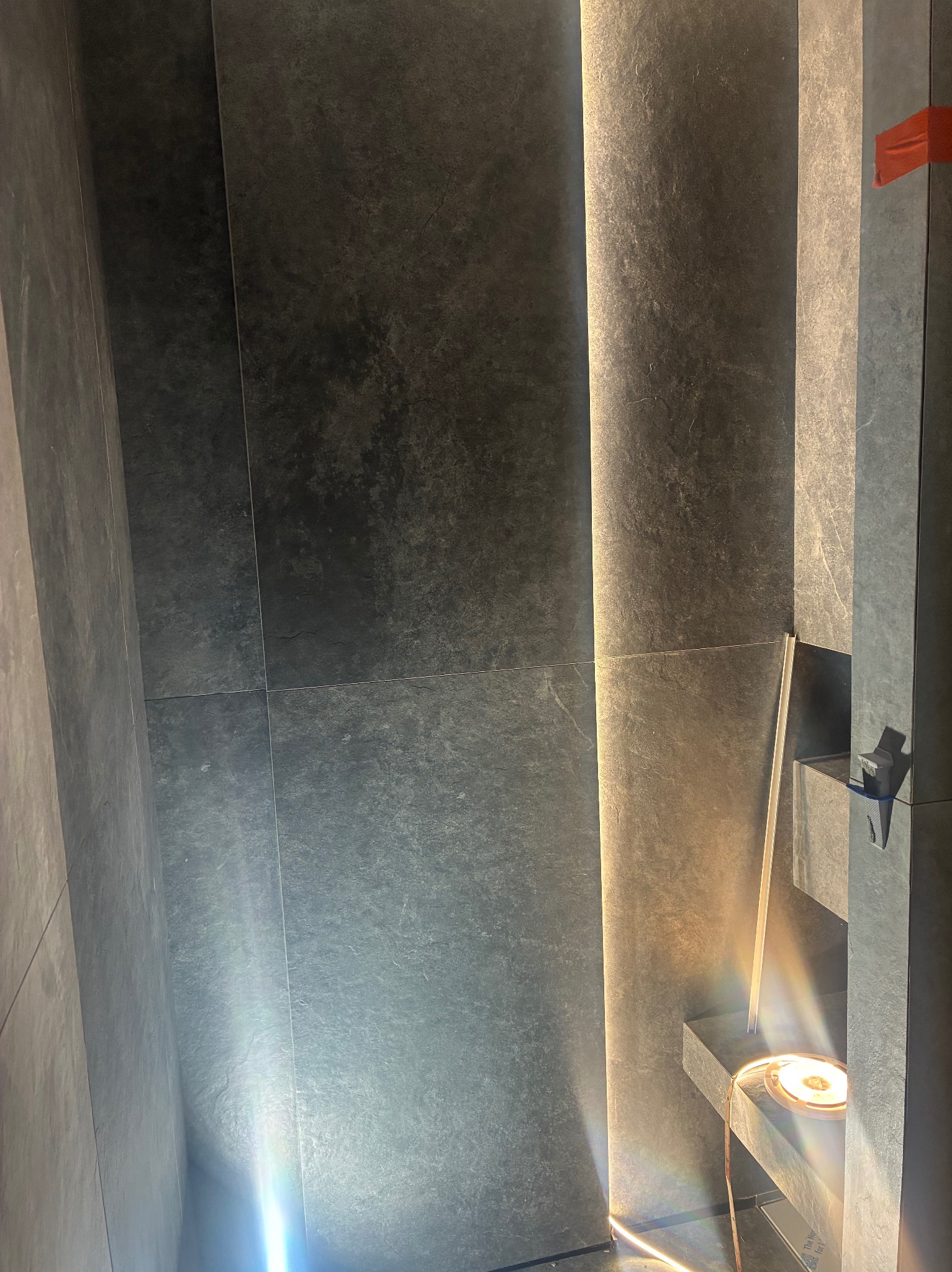 Ciemne, matowe płytki w łazience z podświetleniem LED wzdłuż pionowych krawędzi. Widoczne detale montażowe i poziomica, sugerujące świeżo zakończone prace wykończeniowe.