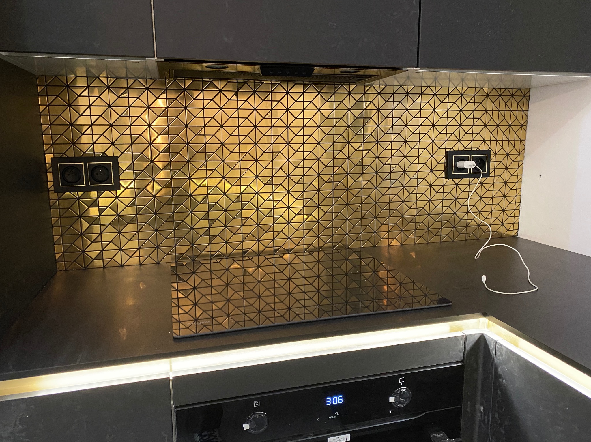 Kuchnia z czarnymi szafkami i złotą mozaiką na ścianie nad blatem. Widoczne gniazdka elektryczne i płyta indukcyjna. Oświetlenie LED pod szafkami dodaje blasku.