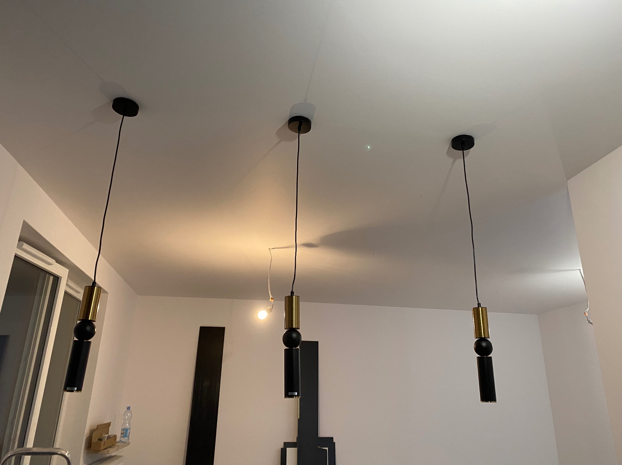 Trzy nowoczesne lampy wiszące z czarnymi i złotymi elementami, równomiernie rozmieszczone na białym suficie w jasnym wnętrzu. Minimalistyczny design oświetlenia.