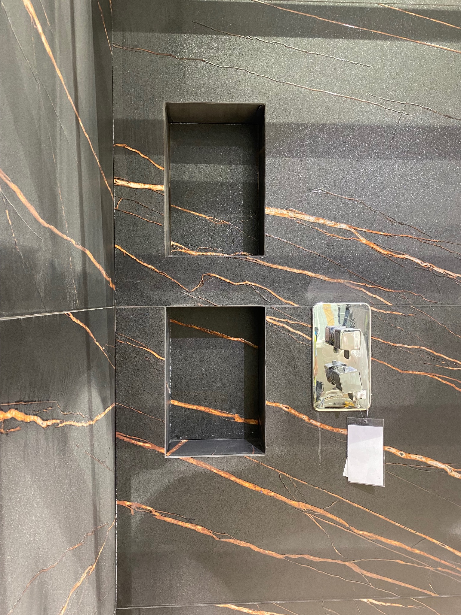 Czarne płytki imitujące marmur z złotymi żyłami w kabinie prysznicowej. W ścianie wnęki, bateria prysznicowa z zawieszką. Wykończenie łazienki w nowoczesnym stylu.