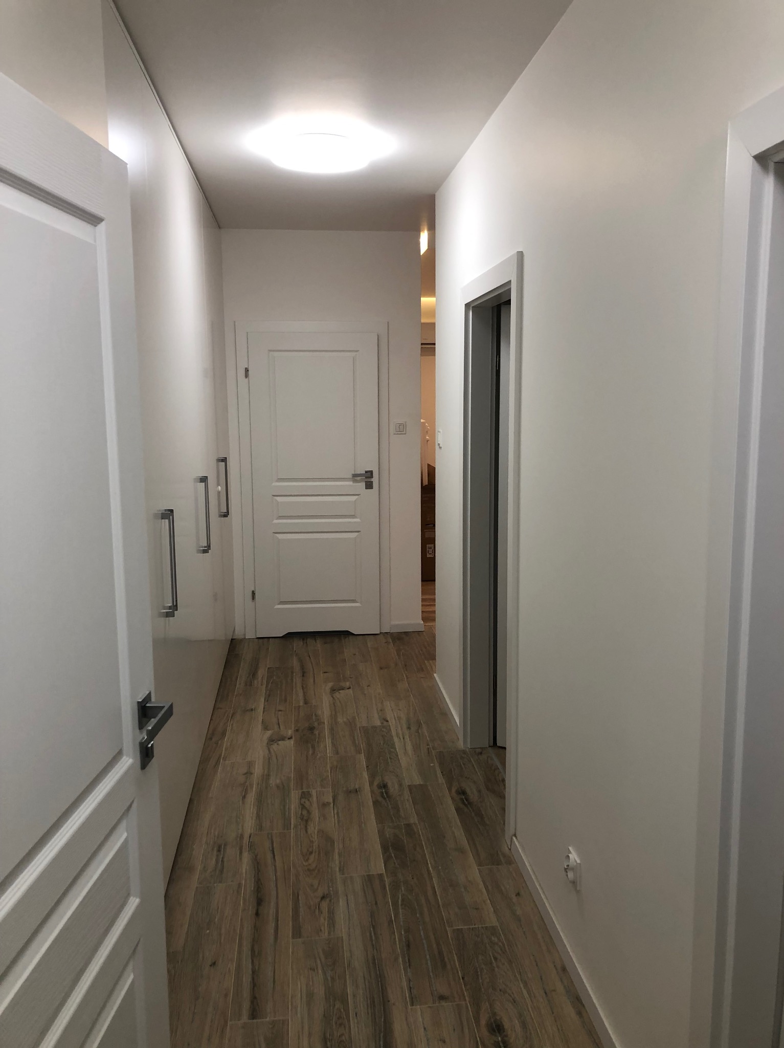 Biały korytarz z drewnopodobną podłogą, zabudowaną szafą i drzwiami. Widoczne detale wykończenia, takie jak listwy przypodłogowe i oświetlenie sufitowe.