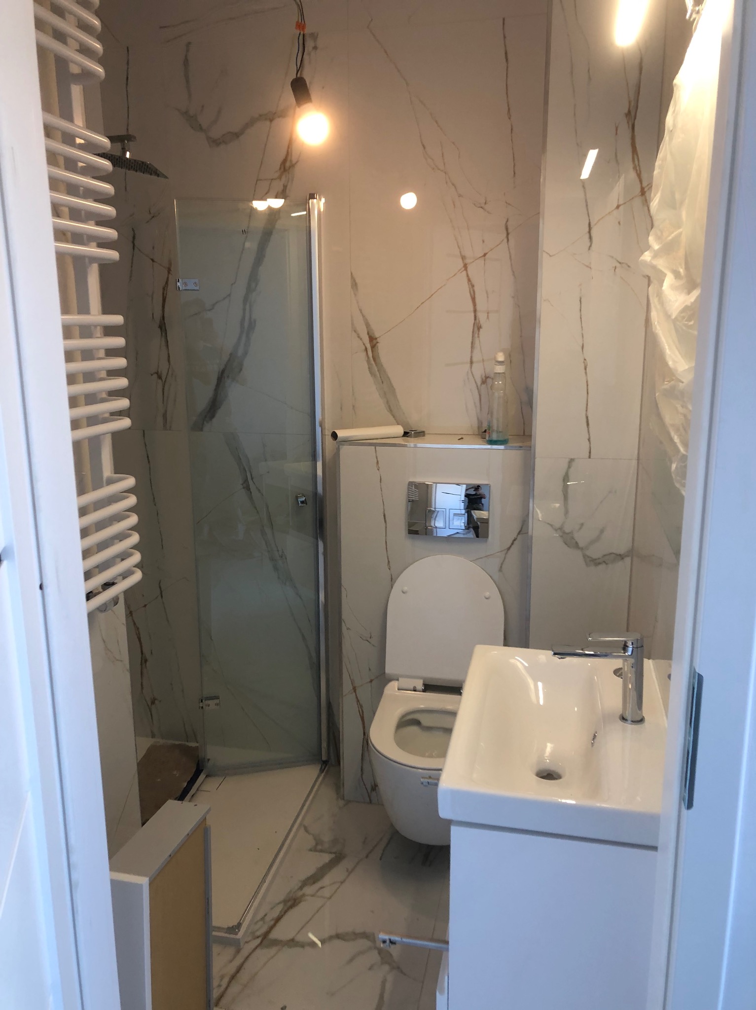 Wykończona łazienka z kabiną prysznicową, toaletą i umywalką w nowoczesnym stylu. Białe płytki imitujące marmur z brązowymi żyłami na ścianach i podłodze.