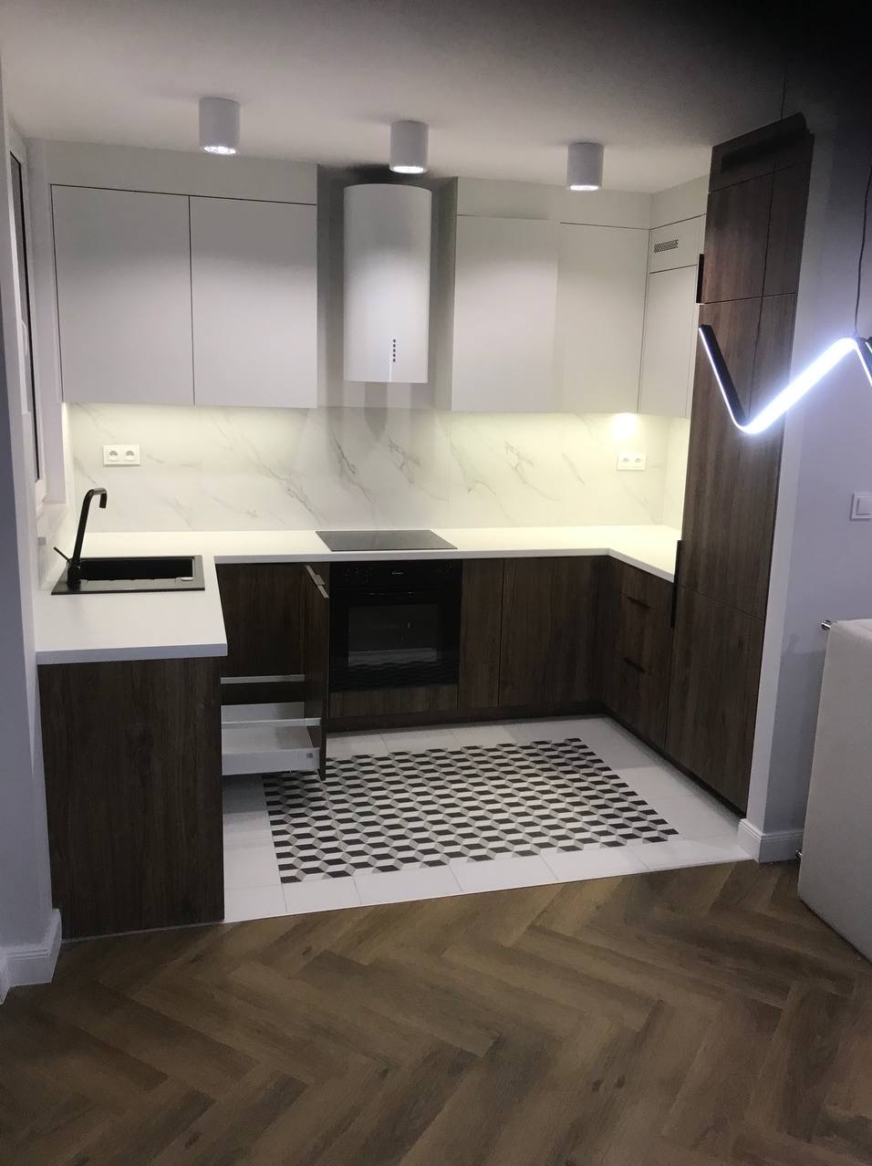 Nowoczesna kuchnia w bieli i drewnie: białe szafki górne, drewniane dolne, wzorzysta podłoga. Otwarta szuflada, minimalistyczny design, oświetlenie punktowe.