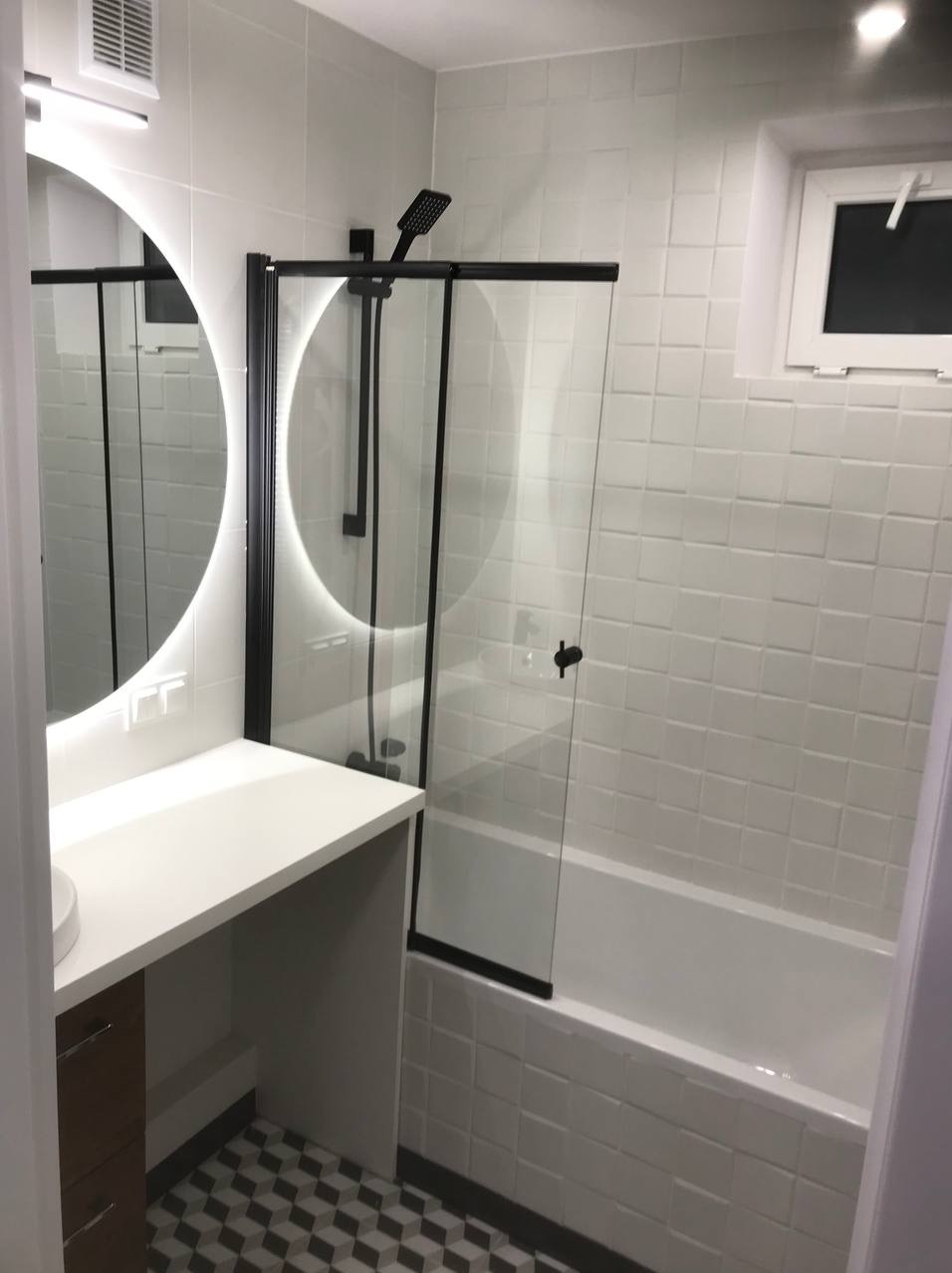 Nowoczesna łazienka z białymi płytkami, kabiną prysznicową z czarnymi ramami i okrągłym lustrem z podświetleniem LED. Umywalka z blatem i szafką. Geometryczna podłoga.