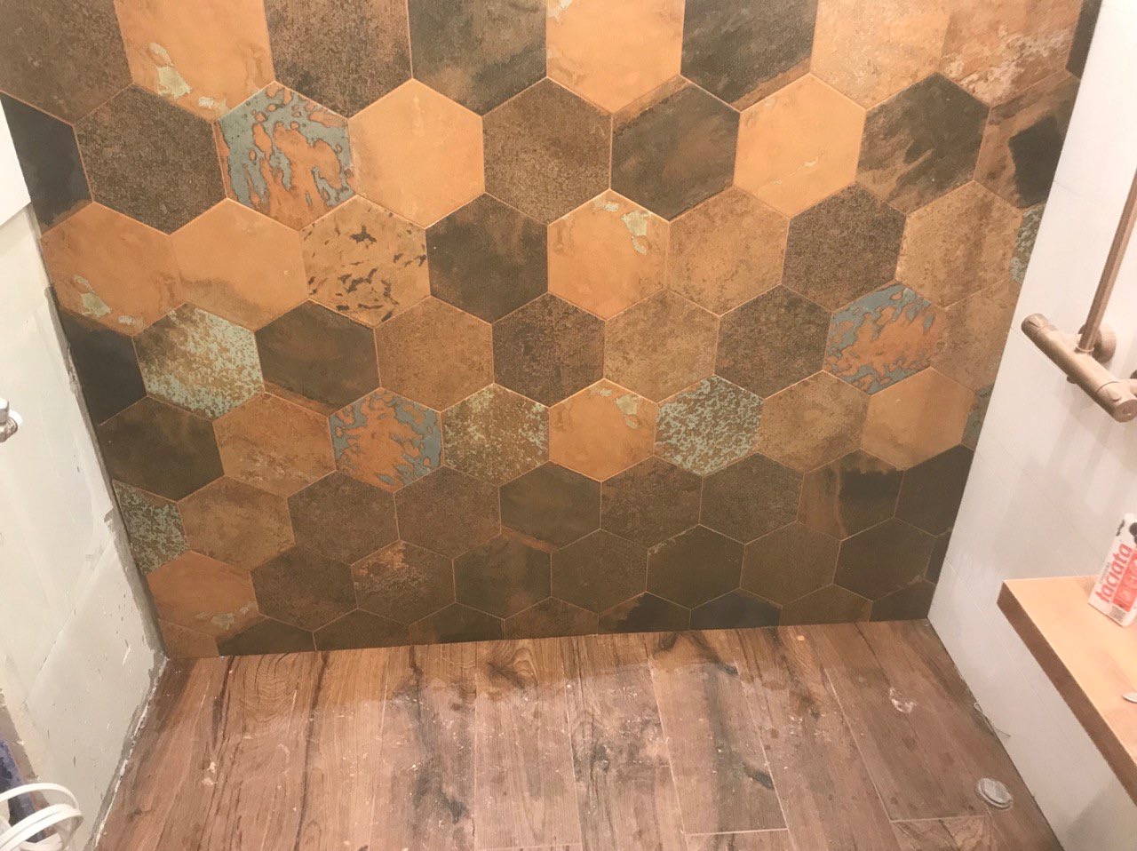 Ściana w łazience wyłożona heksagonalnymi płytkami w odcieniach brązu, miedzi i turkusu, z drewnopodobną podłogą. Widoczny fragment półki i armatury.