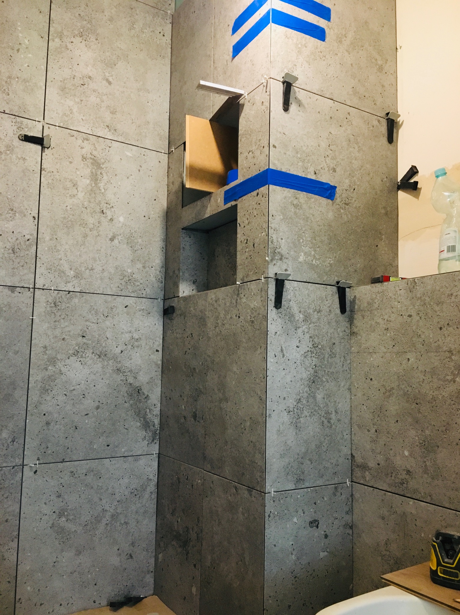 Szare, kwadratowe płytki imitujące kamień, ułożone w łazience, z niebieską taśmą i narzędziami glazurniczymi, tworzą wnękę. Widoczne elementy poziomowania płytek.