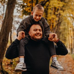 MARCIN SALWIAK FOTOGRAFIA - Uśmiechnięty mężczyzna z brodą niesie na barana uśmiechniętego chłopca w jesiennym lesie. Ciepłe światło, naturalne kolory, widoczne drzewa i opadłe liście.