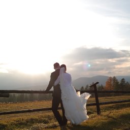 MARCIN SALWIAK FOTOGRAFIA - Para młoda, odwrócona tyłem, opiera się o drewniany płot na tle gór i zachodzącego słońca. Suknia powiewa na wietrze. Romantyczna sceneria.