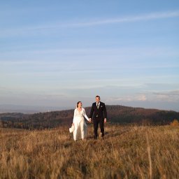 MARCIN SALWIAK FOTOGRAFIA - Para młoda, trzymając się za ręce, spaceruje po łące w jesiennej scenerii. W tle wzgórza i błękitne niebo. Ujęcie z dołu podkreśla przestrzeń i intymność chwili.