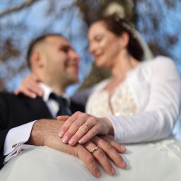 MARCIN SALWIAK FOTOGRAFIA - Ujęcie dłoni pary młodej z obrączkami na tle rozmytej sylwetki nowożeńców i jesiennego krajobrazu. Romantyczna fotografia ślubna z bliska.