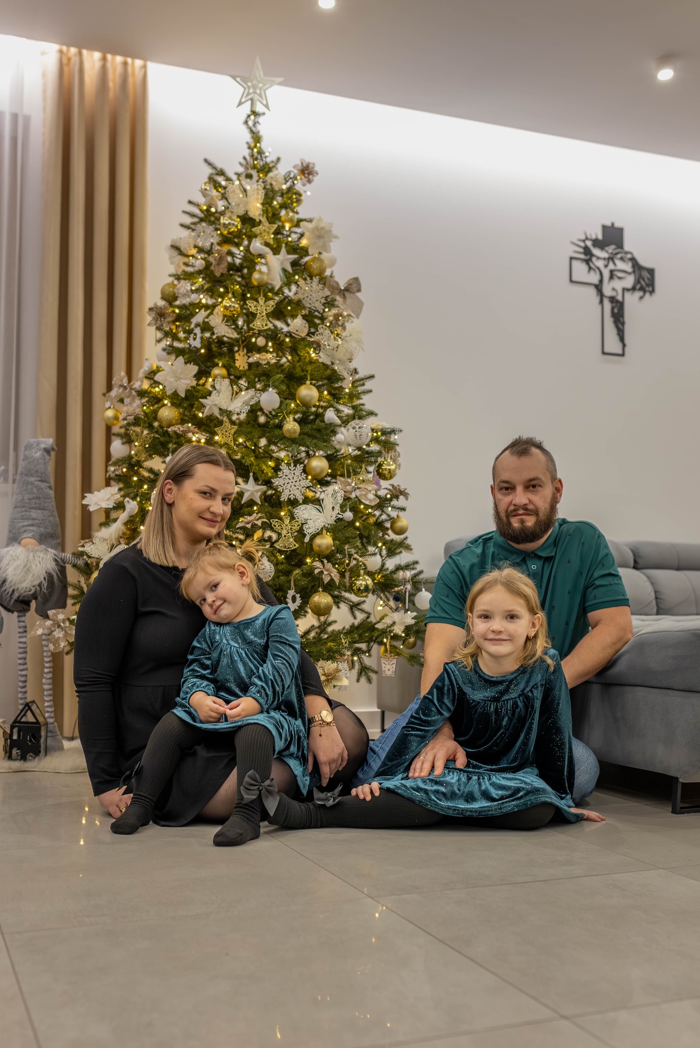 Rodzinne zdjęcie portretowe z dwójką dzieci przed bogato zdobioną choinką w ciepłym, domowym wnętrzu z krzyżem na ścianie.