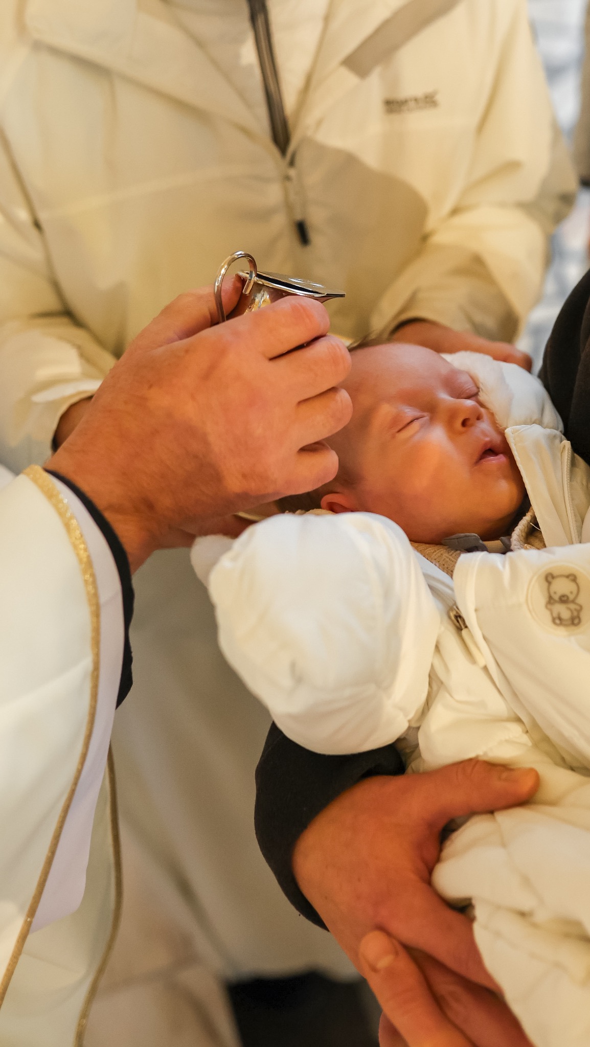 Zbliżenie na moment chrztu: ksiądz polewa wodą główkę śpiącego niemowlęcia ubranego w biały kombinezon z misiem, trzymanego przez rodzica. Widoczne detale szat liturgicznych.