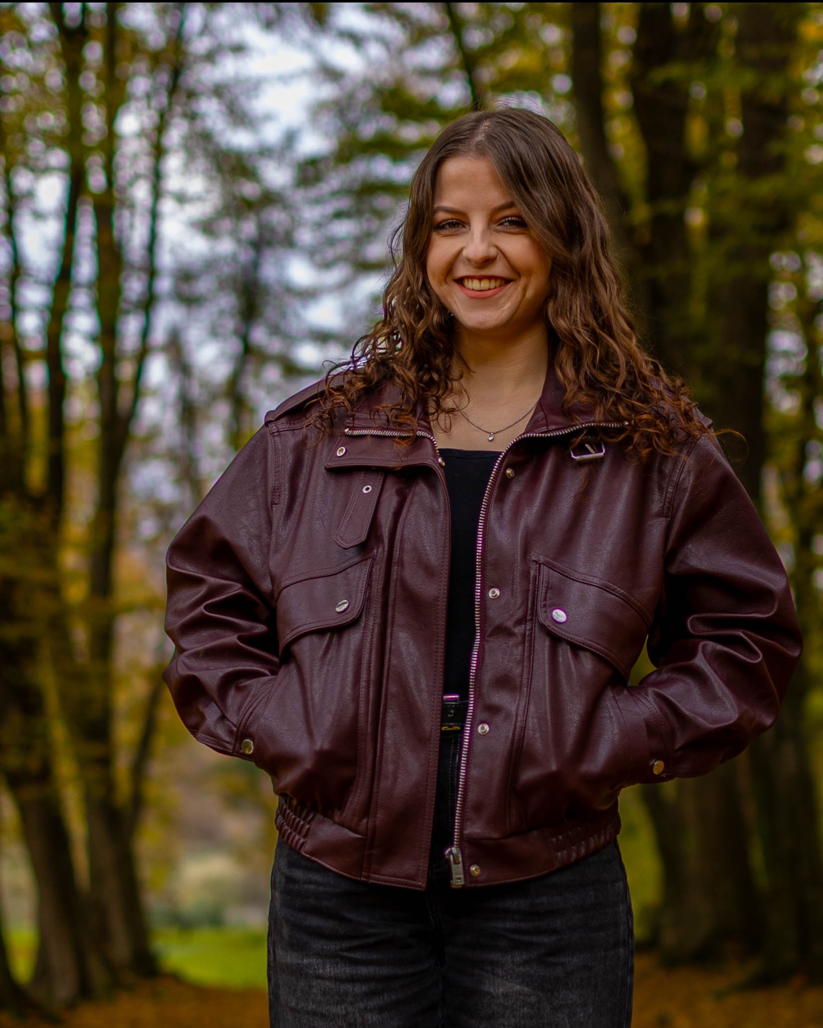 Uśmiechnięta brunetka w bordowej kurtce bomberce, stojąca na tle jesiennego lasu. Swobodna poza, ręce w kieszeniach, naturalne światło.