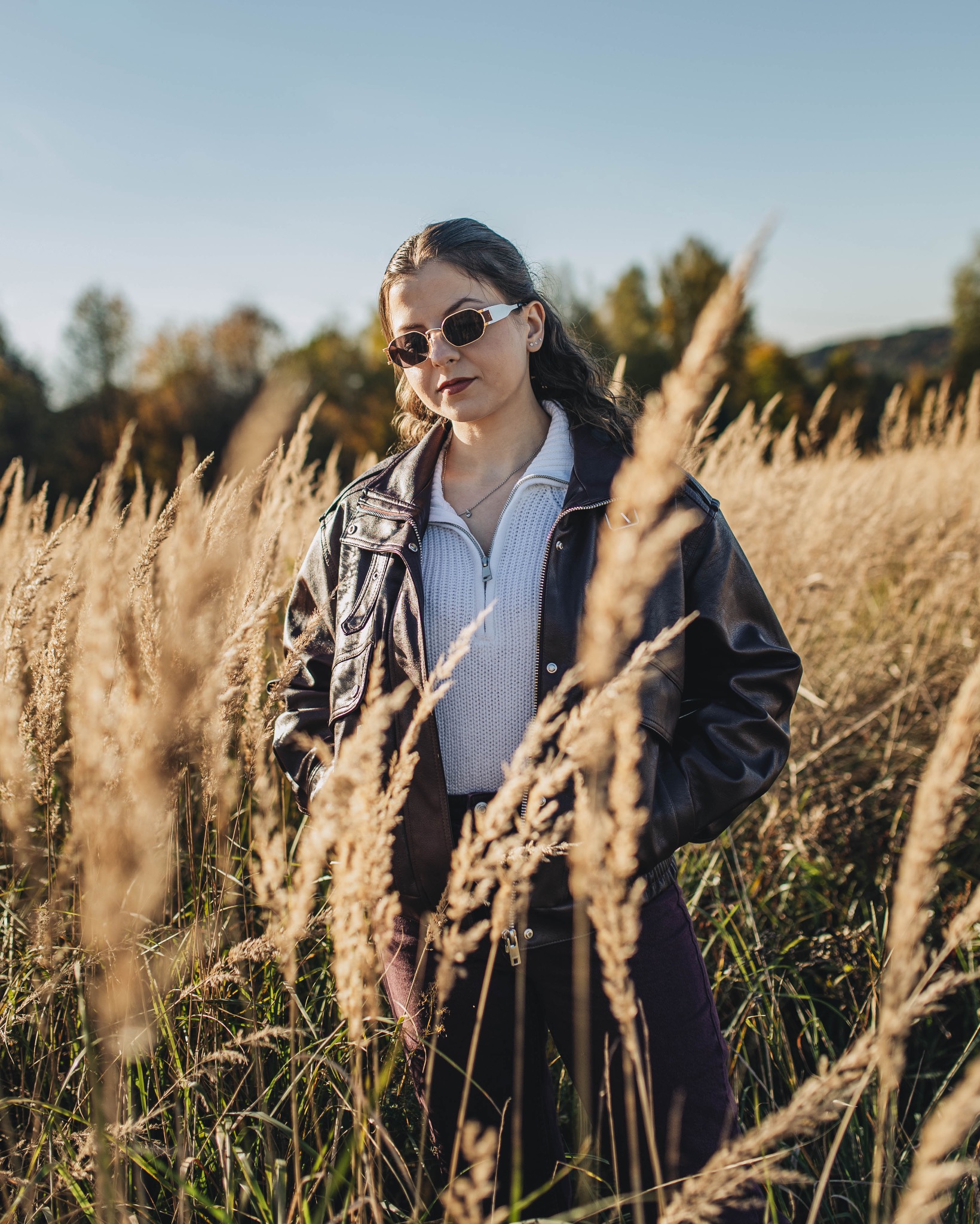 Portret kobiety w okularach przeciwsłonecznych, w brązowej kurtce, stojącej na tle pola trzcin w słoneczny dzień. Ujęcie z dołu, z widocznymi drzewami w tle.