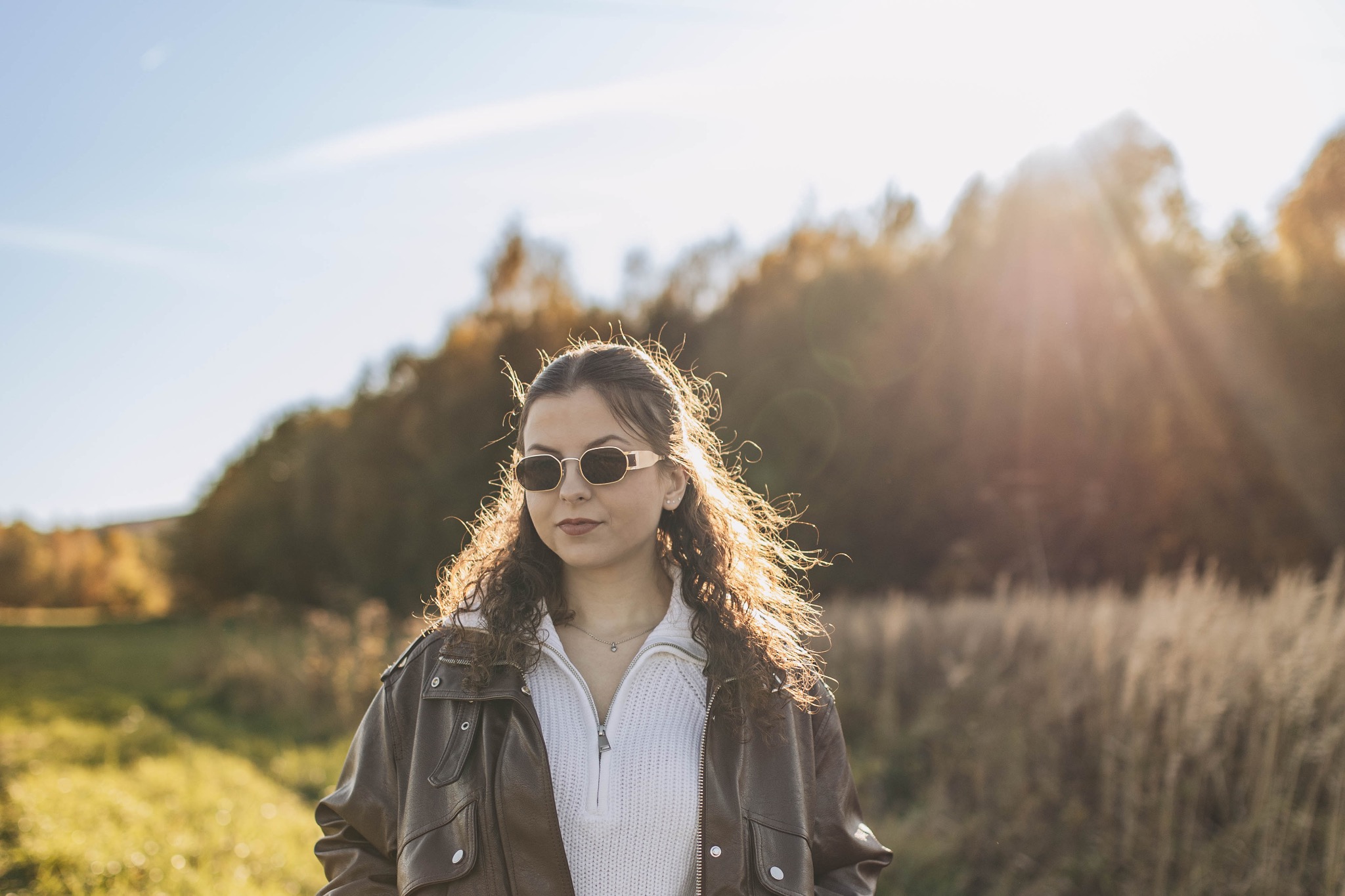 Portret młodej kobiety w okularach przeciwsłonecznych, w brązowej skórzanej kurtce i białym swetrze na tle jesiennego krajobrazu z promieniami słońca.