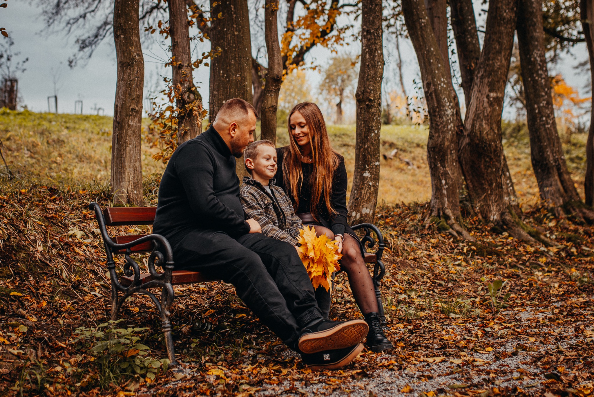 Jesienna sesja rodzinna: rodzice z synem siedzą na ławce w parku, trzymając bukiet żółtych liści. Ciepłe barwy jesieni, naturalne światło, swobodna atmosfera.