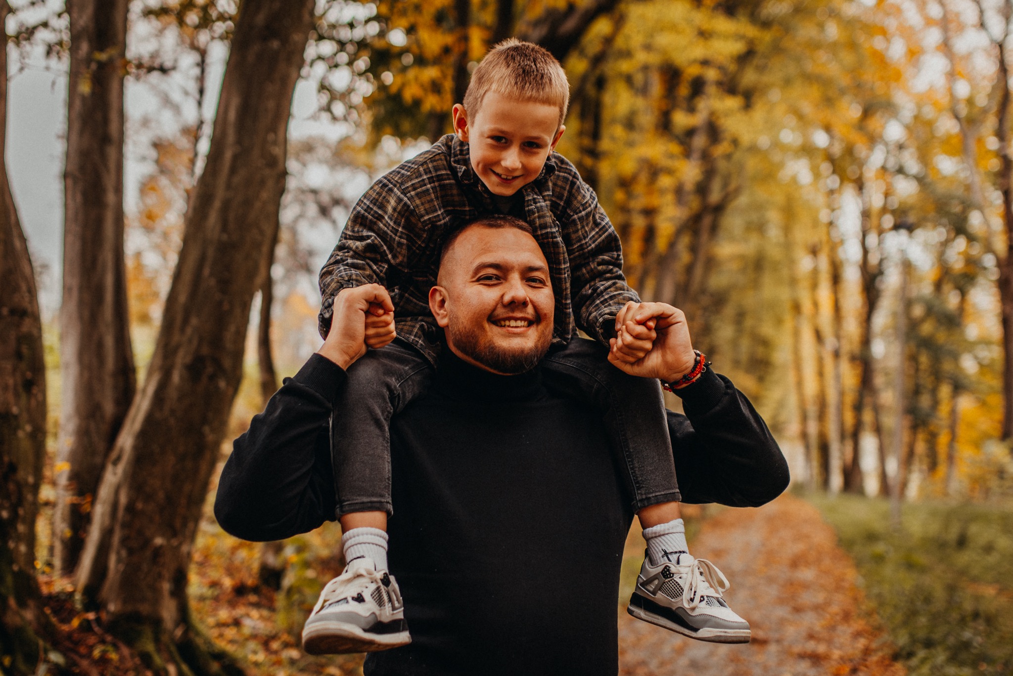 Uśmiechnięty mężczyzna z brodą niesie na barana uśmiechniętego chłopca w jesiennym lesie. Ciepłe światło, naturalne kolory, widoczne drzewa i opadłe liście.