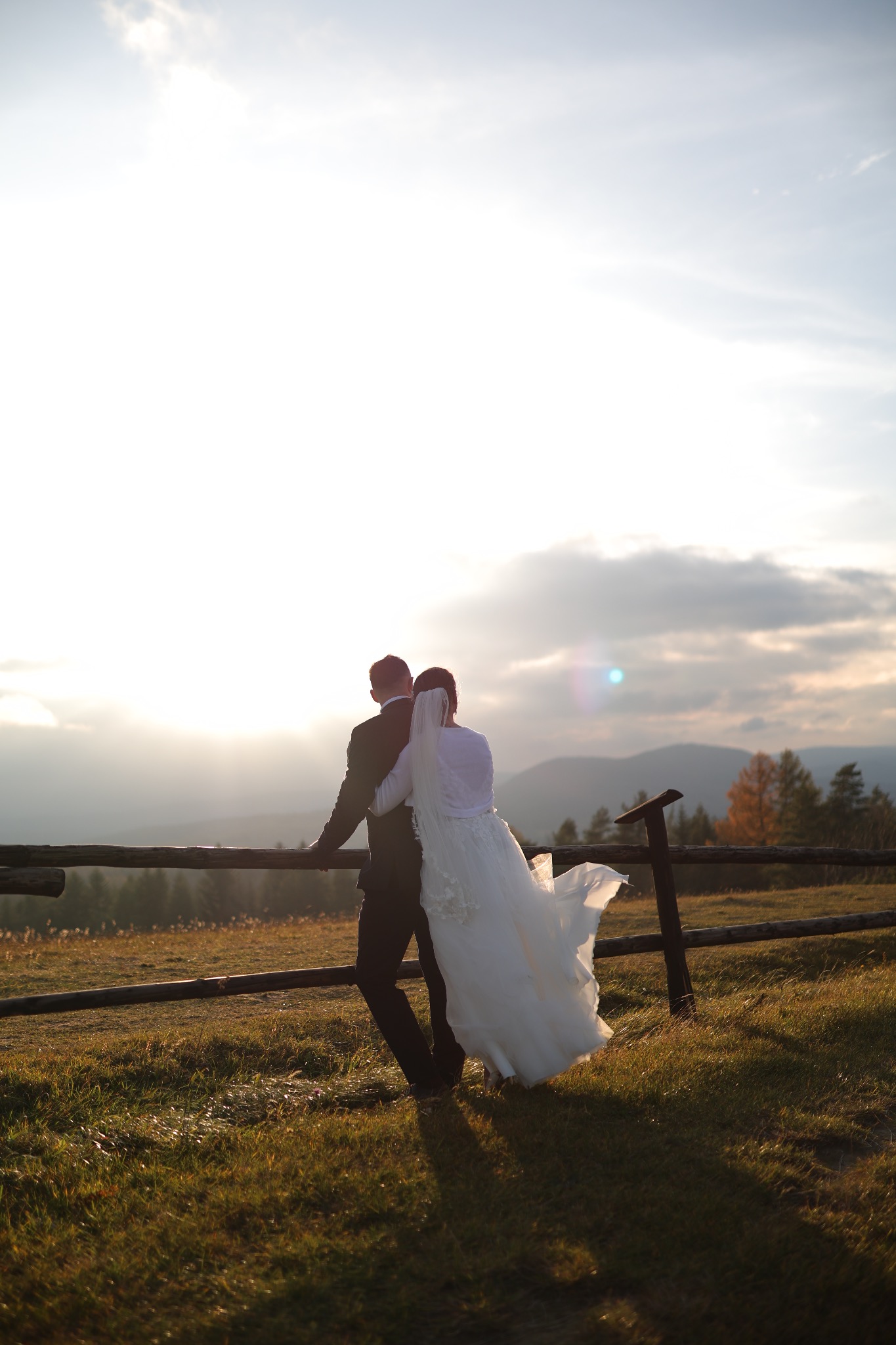 Para młoda, odwrócona tyłem, opiera się o drewniany płot na tle gór i zachodzącego słońca. Suknia powiewa na wietrze. Romantyczna sceneria.