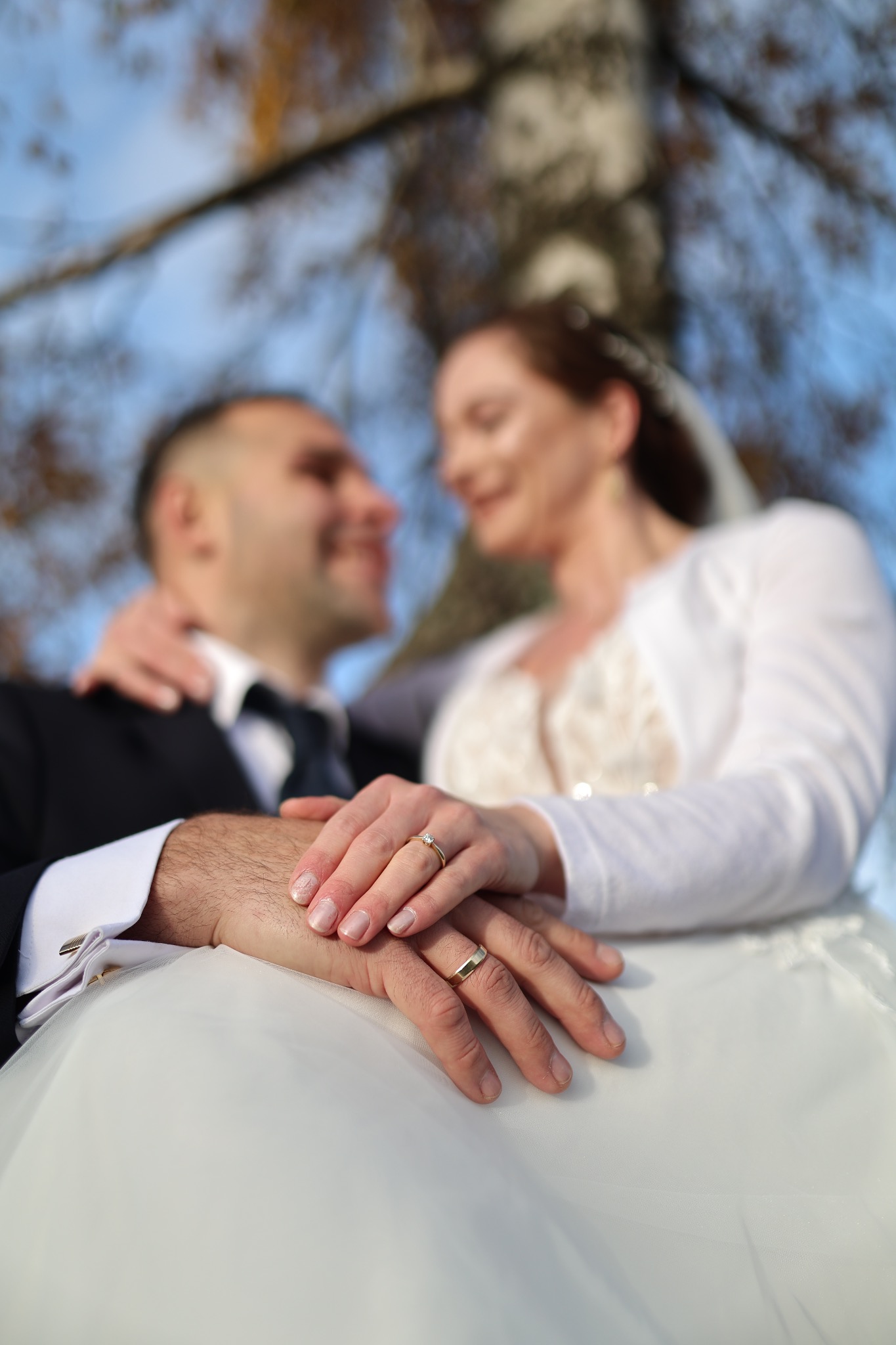 Ujęcie dłoni pary młodej z obrączkami na tle rozmytej sylwetki nowożeńców i jesiennego krajobrazu. Romantyczna fotografia ślubna z bliska.