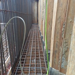 Hydrostop - Hydroizolacje krystalizujące - Zbrojenie fundamentów w wykopie z deskowaniem stalowym i zielonym przewodem. Widoczne pręty pionowe i poziome tworzące siatkę. Etap przygotowania do zalewania betonem.