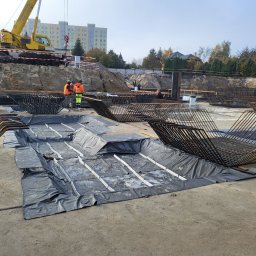 Hydrostop - Hydroizolacje krystalizujące - Budowa fundamentów: zbrojenie, izolacja i pracownicy w pomarańczowych kamizelkach na placu budowy z dźwigiem w tle. Widok na przygotowanie pod wylewkę.